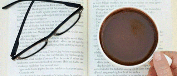 Someone holding a cup of tea with a reading glass and a book