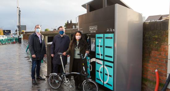 People standing with a foldable bike hired from a Brompton Bike Hire machine behind them