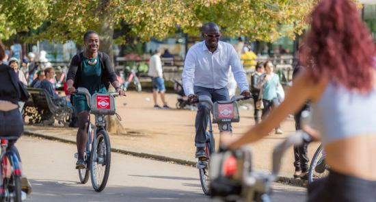 A man and woman riding a Santander Cycle bike through a busy London park.
