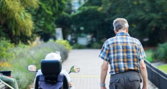 An older couple going along a path together, the gentleman walking and the woman using an ECV