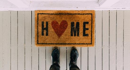 Person standing in front of their frontdoor by the floor mat. The floor mat says home on it with a heart.